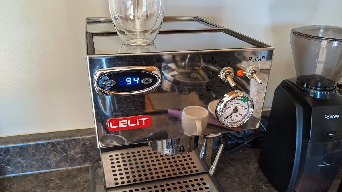 Steel semi-automatic espresso machine with a pressure gauge and temperature display next to a grinder