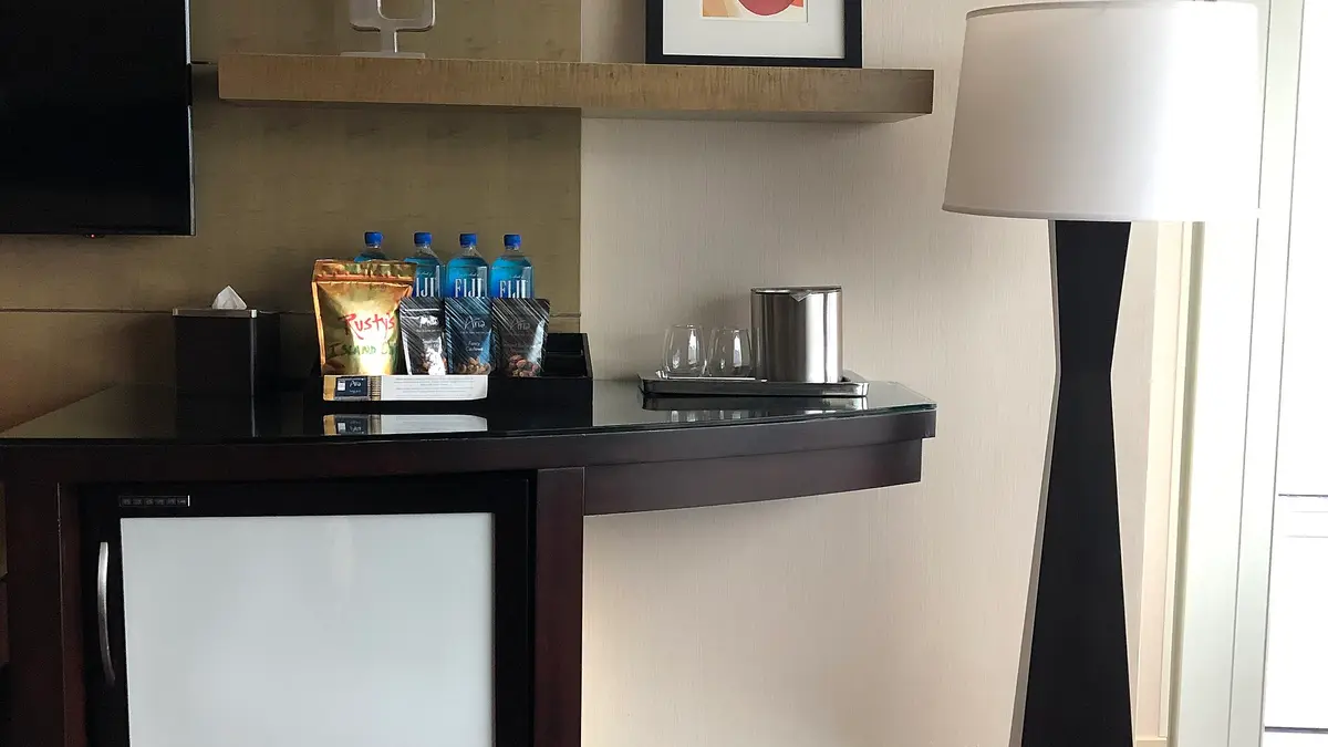 Quarto de hotel com um frigobar branco sob um móvel e garrafas de água em cima
