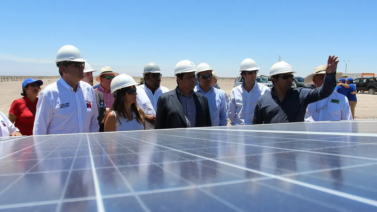 Visitantes con cascos blancos junto a una fila de paneles solares en la planta Pozo Almonte